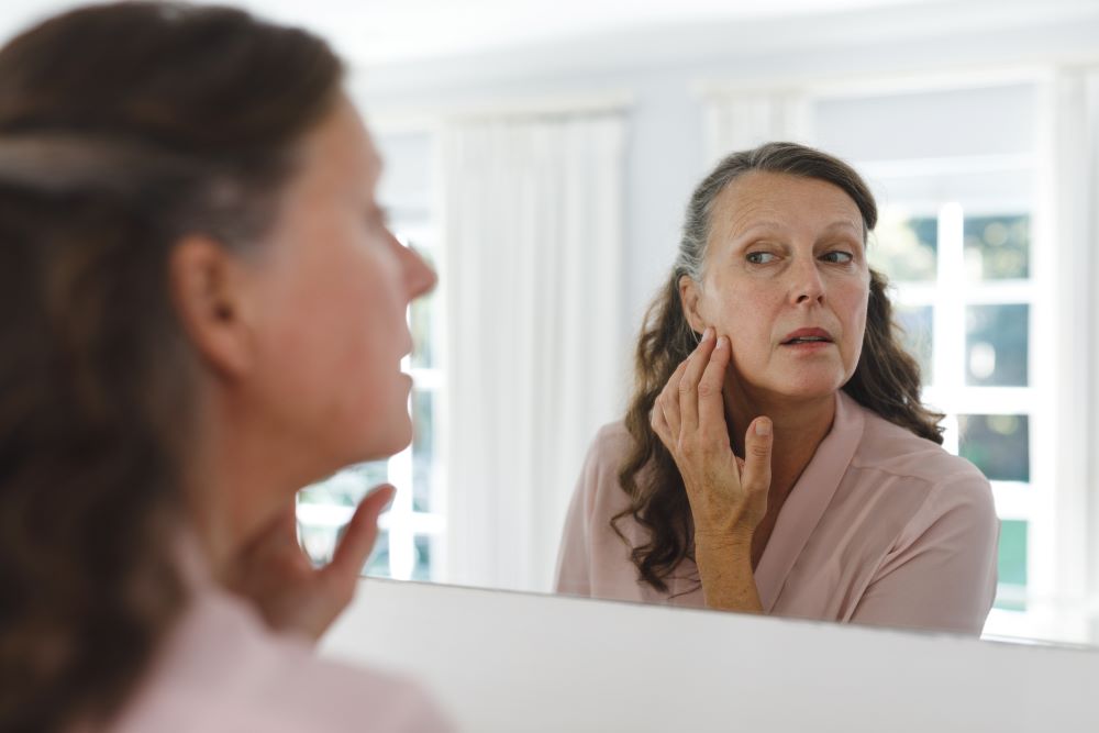 Mulher observa o próprio reflexo no espelho enquanto avalia a região da mandíbula e das bochechas, ilustrando a perda de definição do contorno facial e o interesse por tratamentos de rejuvenescimento.