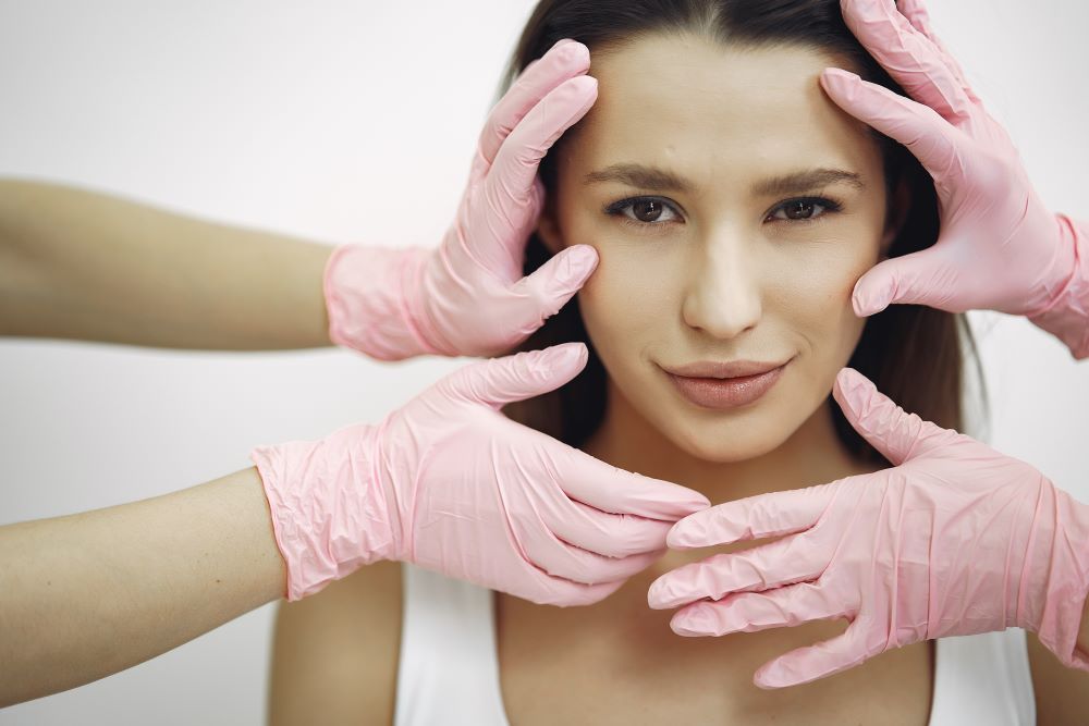 Mulher jovem em ambiente de consultório médico, com o rosto sendo delicadamente examinado por mãos de profissional de saúde com luvas cirúrgicas rosa, evidenciando avaliação facial pré-procedimento estético para análise de contornos do rosto, mandíbula e simetria.