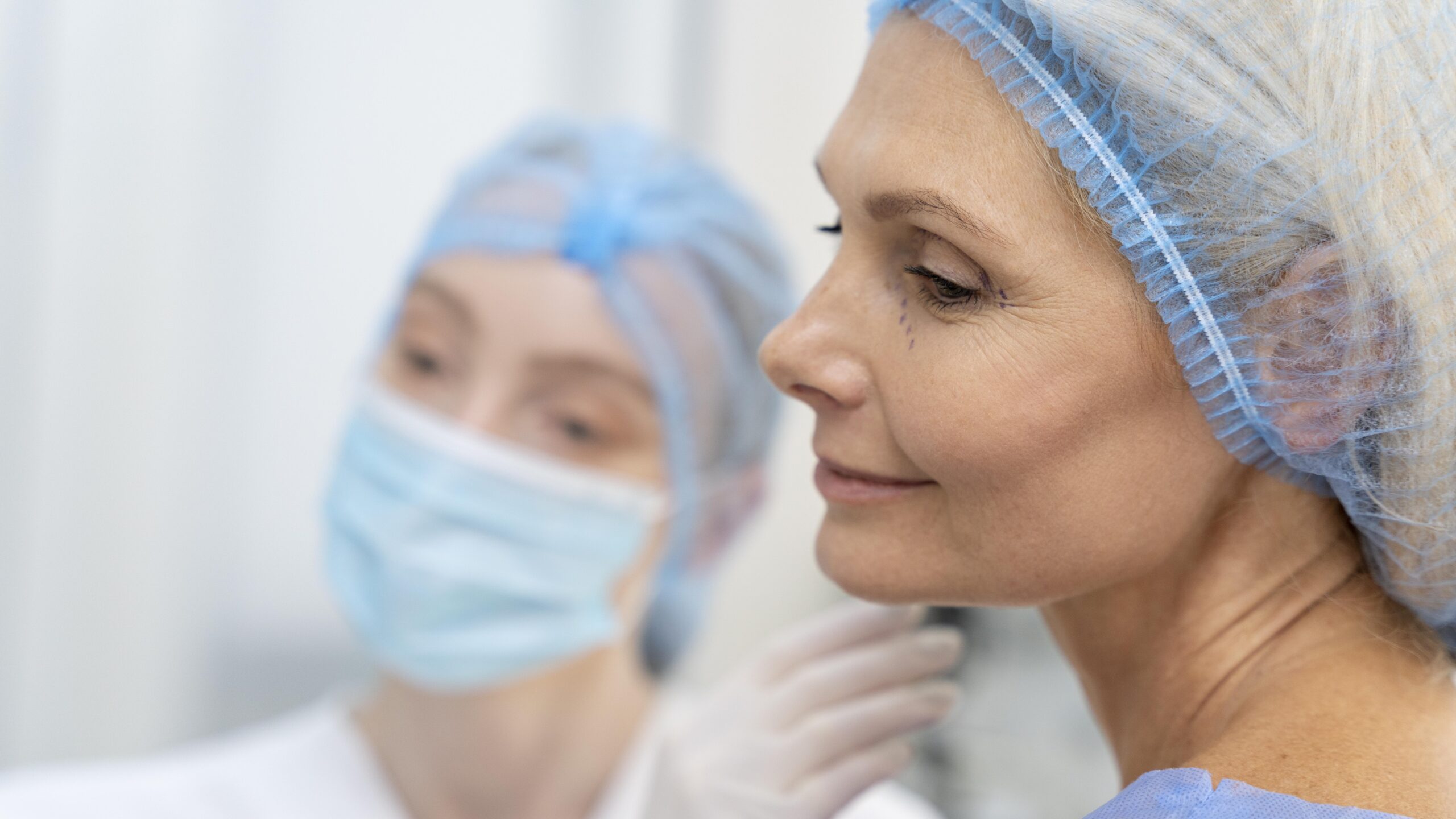 Cirurgião plástico com máscara e luvas examina o rosto de paciente madura com marcações cirúrgicas próximas aos olhos, em preparação para procedimento de rejuvenescimento facial.